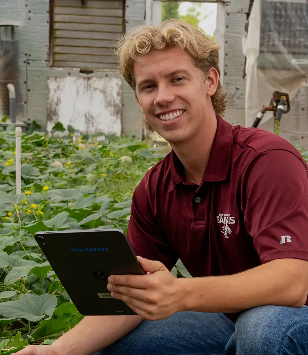 Student in greenhouse reviewing crop plants while holding a tablet.
