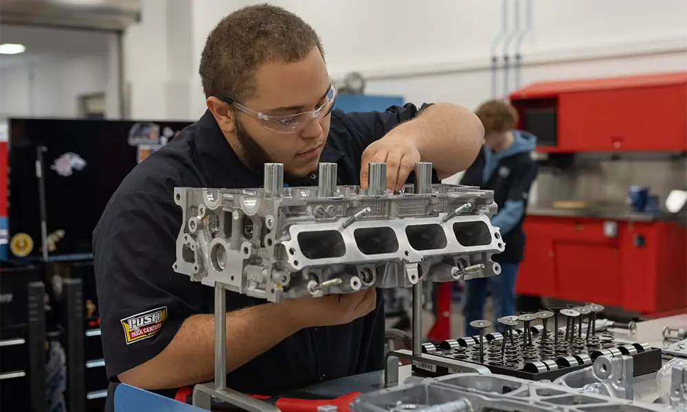 A student wearing safety glasses works carefully on an automotive engine component in a lab, examining and assembling parts on a workbench with tools and engine valves nearby.