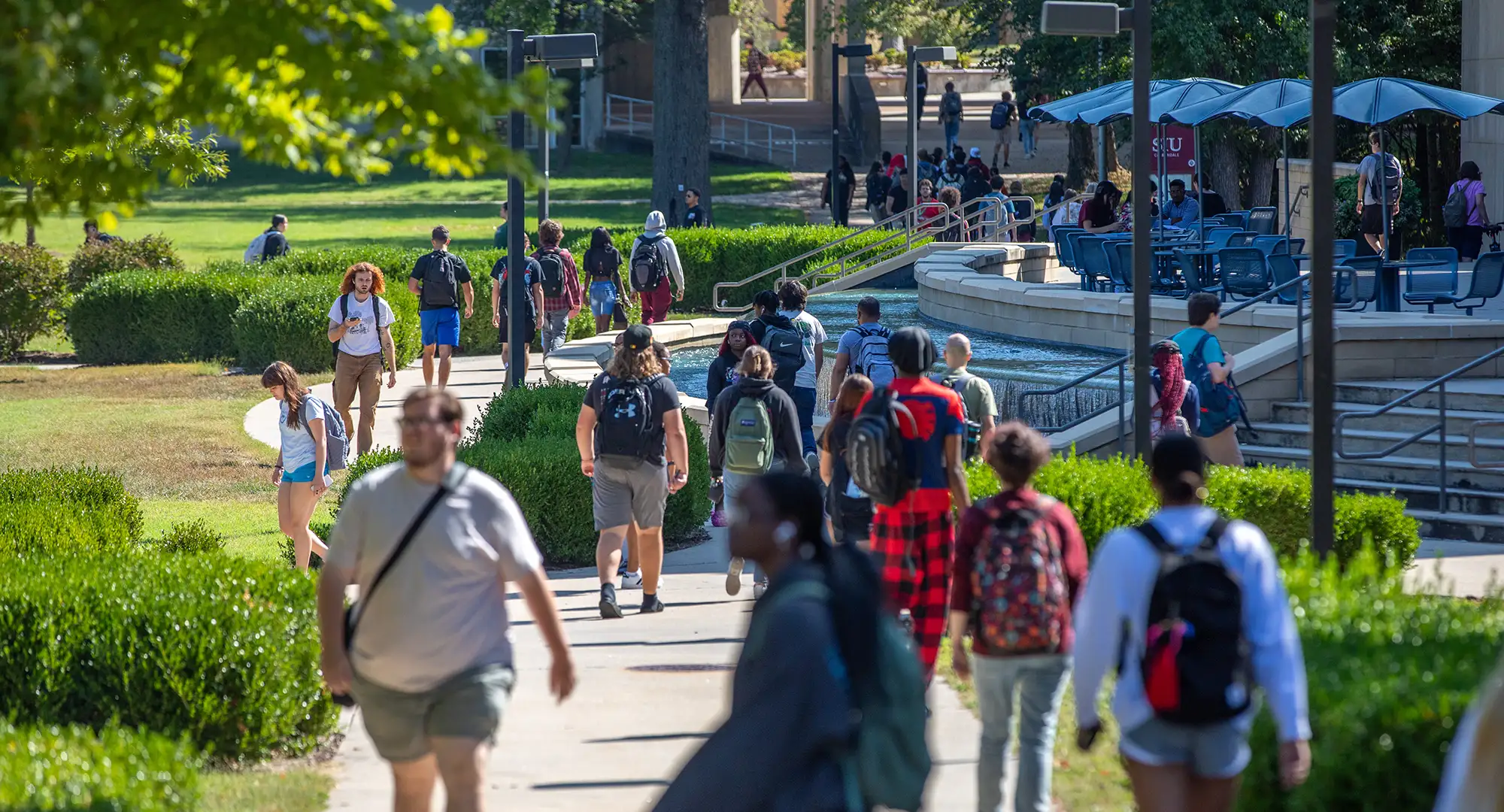 Students walk along a busy campus pathway lined with greenery and seating areas on a sunny day, moving between classes and gathering near outdoor tables.