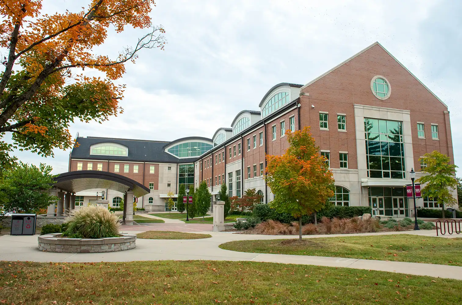 Brick campus building with large windows and arched rooflines surrounded by trees with fall foliage and landscaped walkways.