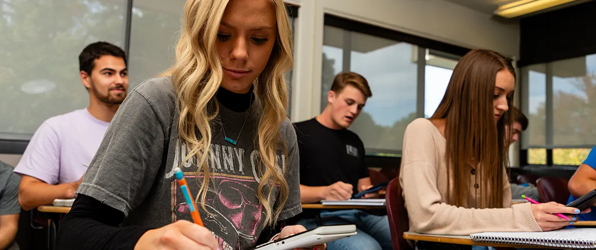 Female college student writing in a notebook while other students take notes in a classroom setting.