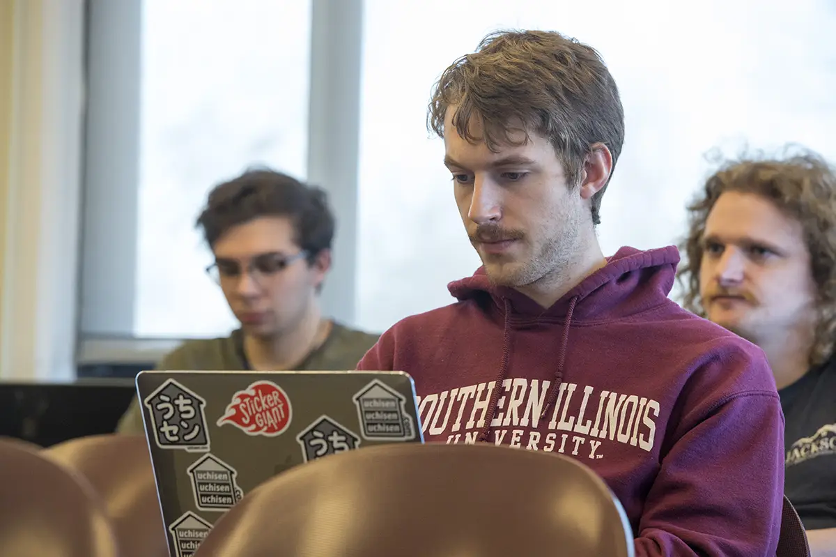 A student in a Southern Illinois University hoodie works on a sticker-covered laptop in a classroom, with two other students visible in the background.