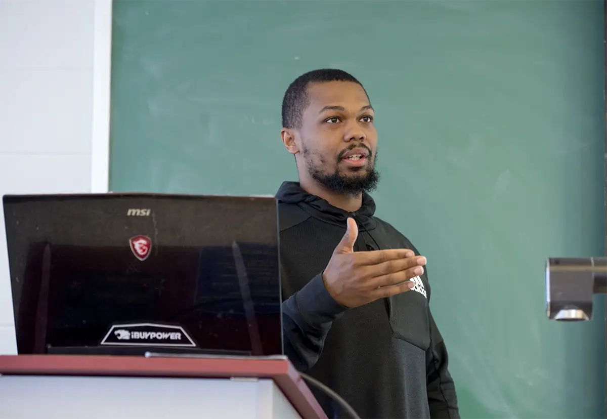 A student presents at a podium with a laptop in a classroom setting.