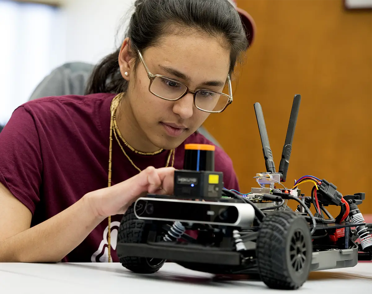 A student in a maroon SIU t-shirt adjusts sensors and components on a small autonomous robotic vehicle in an engineering lab.
