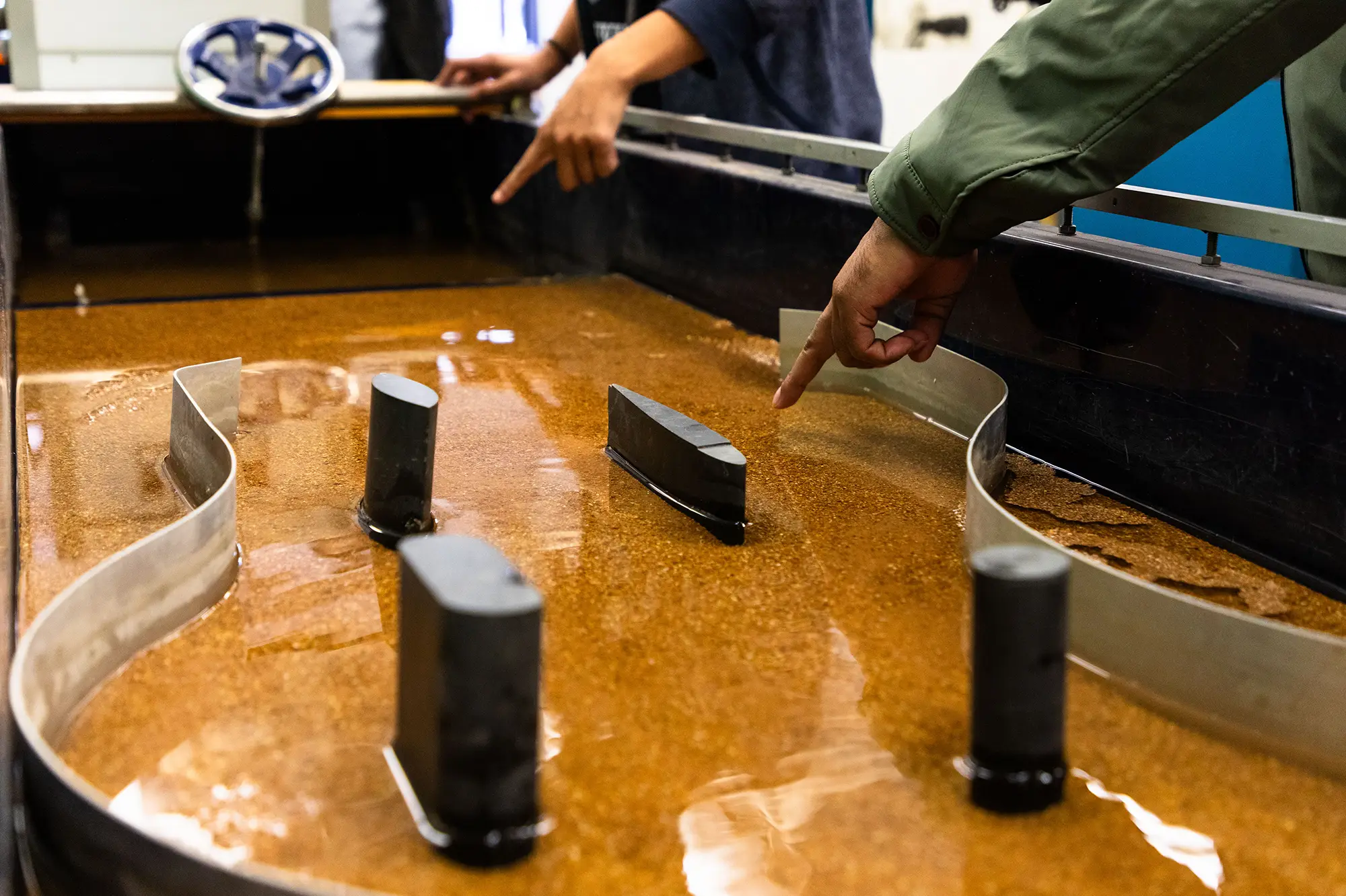 A close-up of a hydraulic engineering research flume with sediment and structural models, with two people pointing at elements in the water flow apparatus.