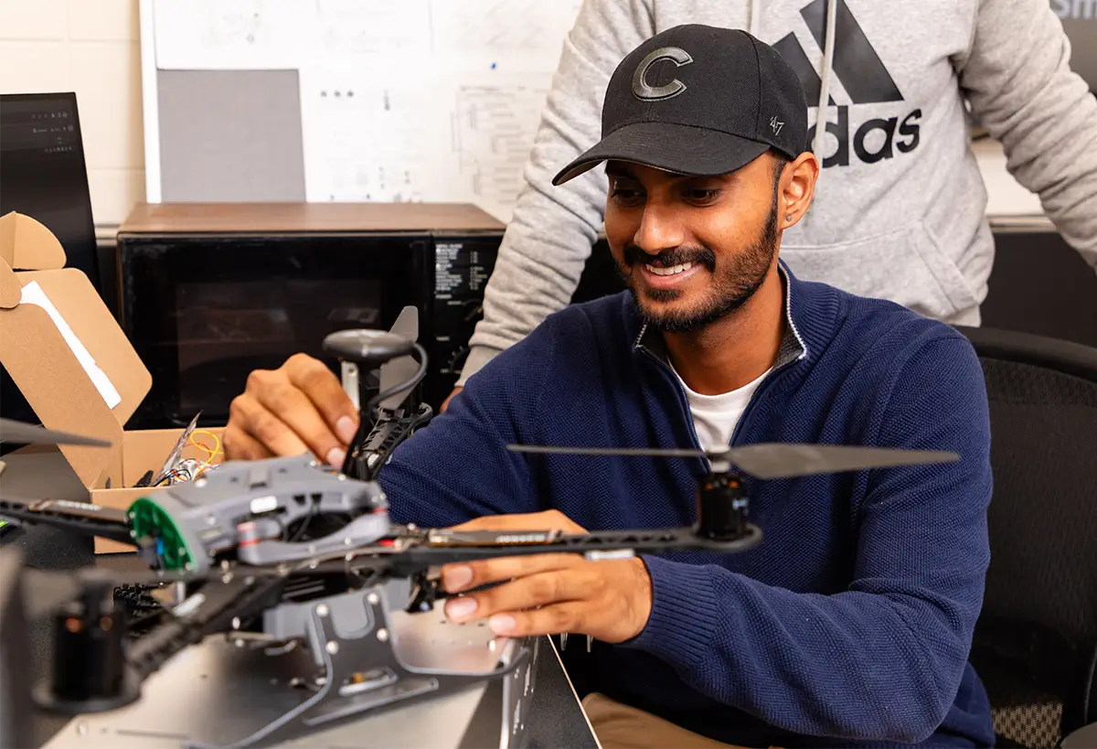 A student assembles or repairs a drone in an engineering lab, with another student visible in the background.