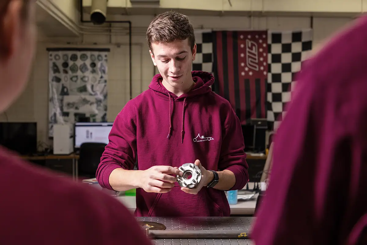 A student in a maroon hoodie examines a machined metal component in an engineering shop, with an SIU flag and checkered banner visible in the background.