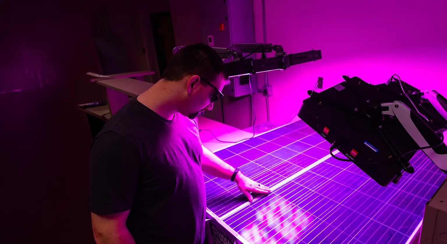 A student wearing safety glasses examines a solar panel under UV lighting in a research laboratory, with a robotic arm visible nearby.