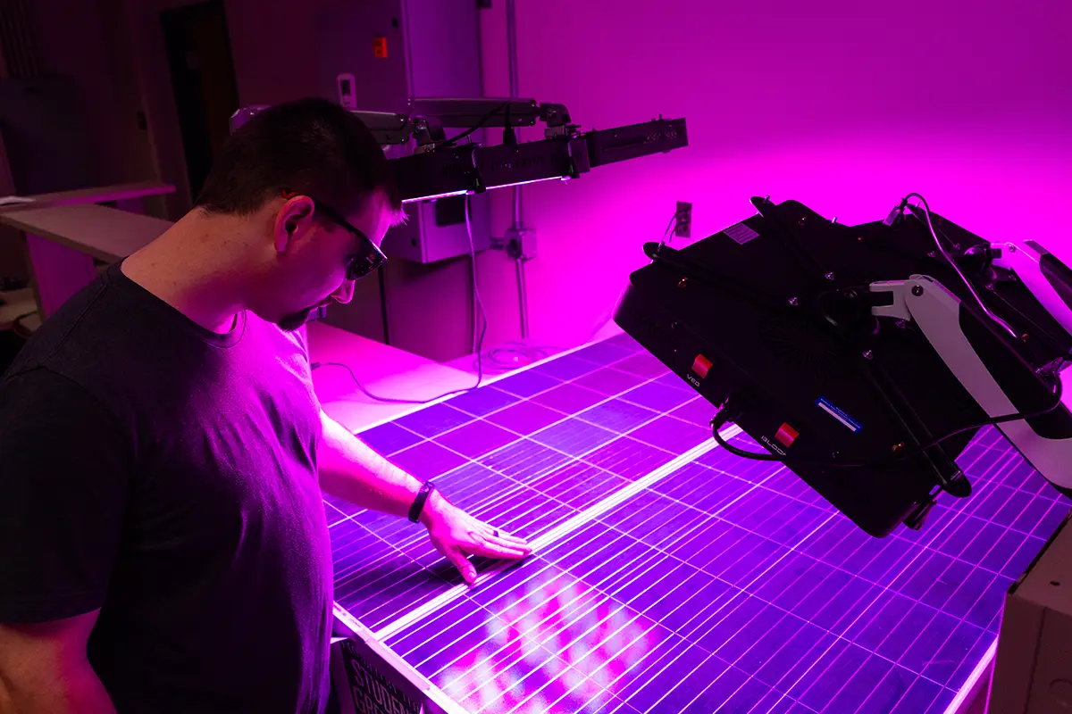 A student wearing safety glasses examines a solar panel under UV lighting in a research laboratory, with a robotic arm visible nearby.
