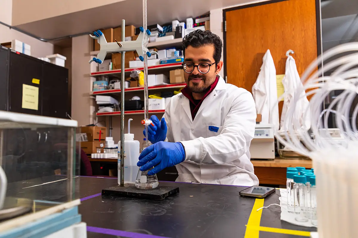 A student in a white lab coat and blue gloves performs a titration experiment with a burette and flask in a chemistry laboratory.