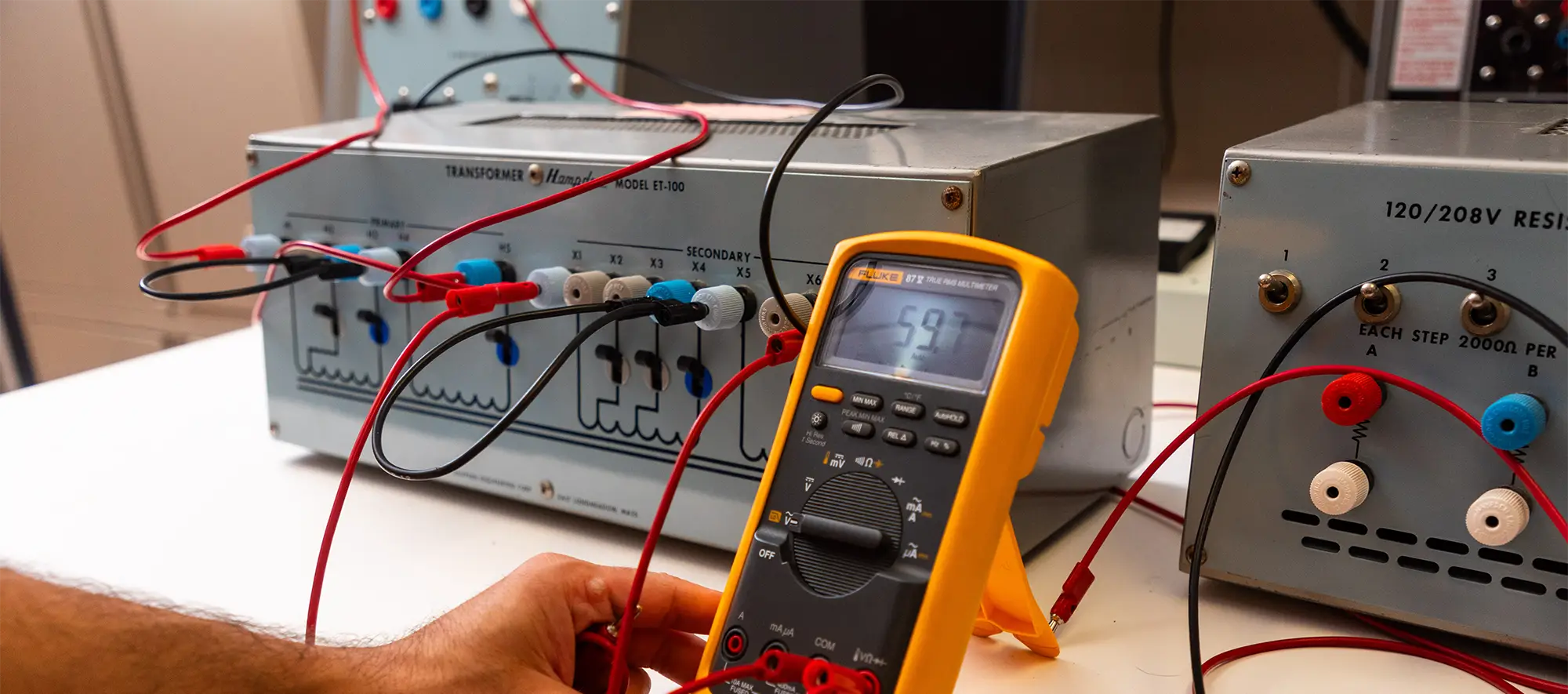 A student uses a Fluke digital multimeter to take measurements on a Hampden transformer training unit in an electrical engineering laboratory.