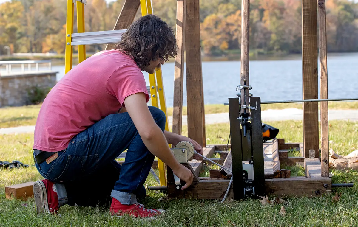 A student adjusts a mechanical engineering apparatus outdoors near a lake on a fall day.