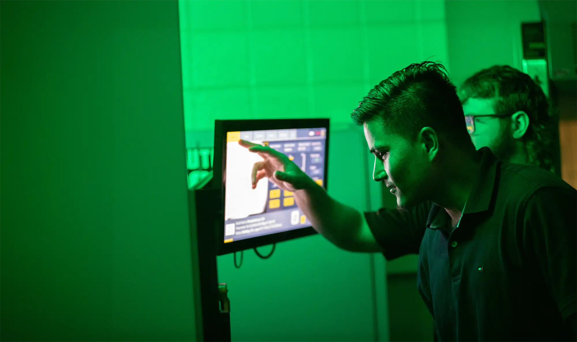 A student interacts with a touchscreen control panel in a green-lit laboratory environment, with a second student visible in the background.