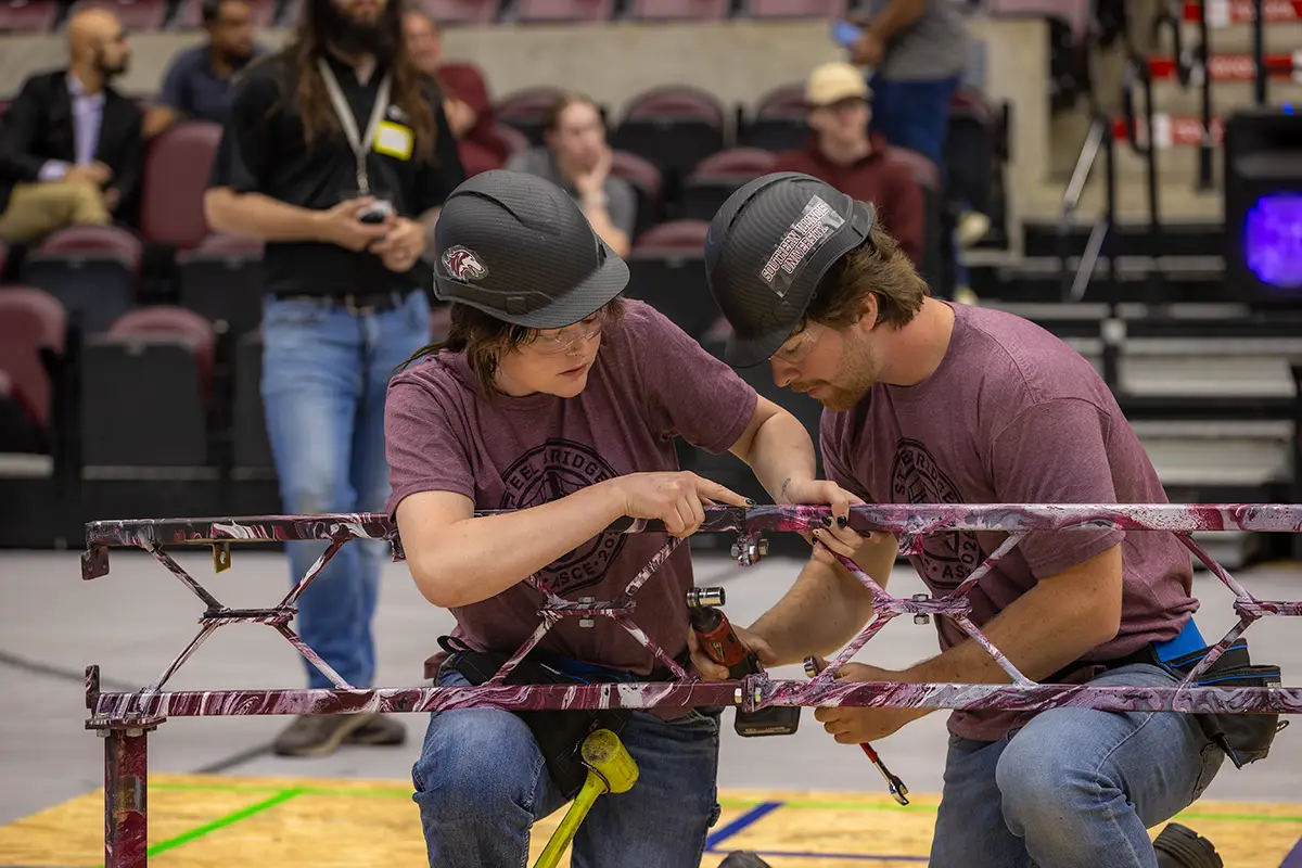 Two SIU Steel Bridge competition team members in hard hats assemble a steel truss structure on an arena floor during a collegiate engineering competition.