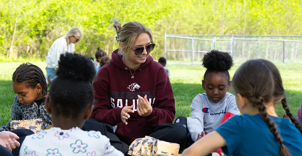 A young woman in a maroon SIU Salukis hoodie sits on grass with a group of young children outdoors, with another adult and additional children visible in the background.