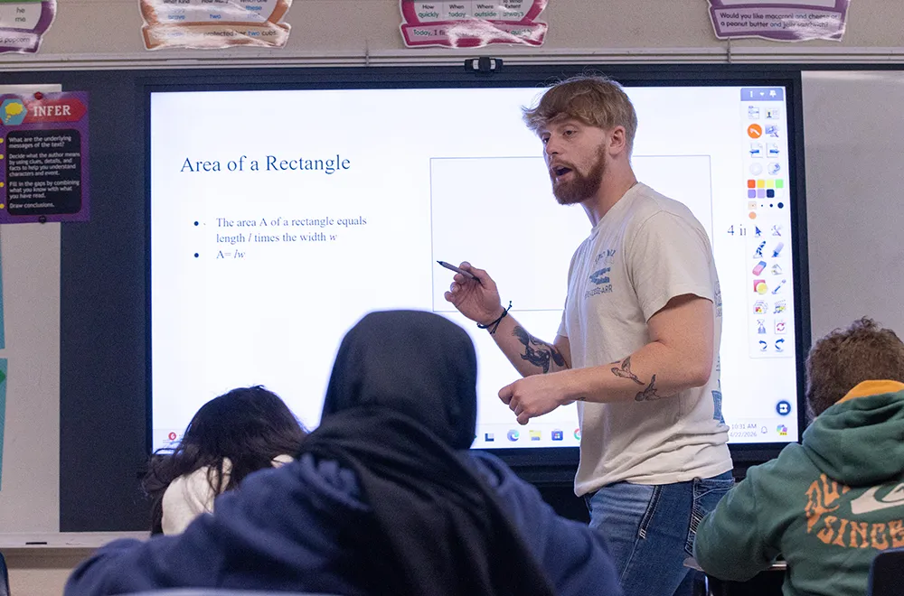 A young man stands at the front of an elementary school classroom presenting a math lesson on area of a rectangle using an interactive whiteboard, with students visible in the foreground.