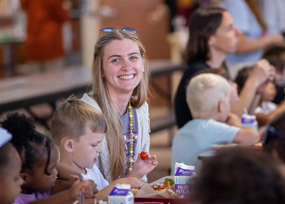 A young woman sits at a cafeteria table eating lunch alongside several young children, with additional adults and children visible in the background.