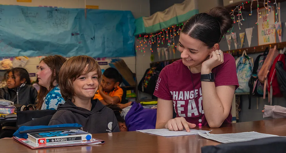 SIU student sits beside an elementary school student at a classroom desk, with other students and classroom decorations visible in the background.