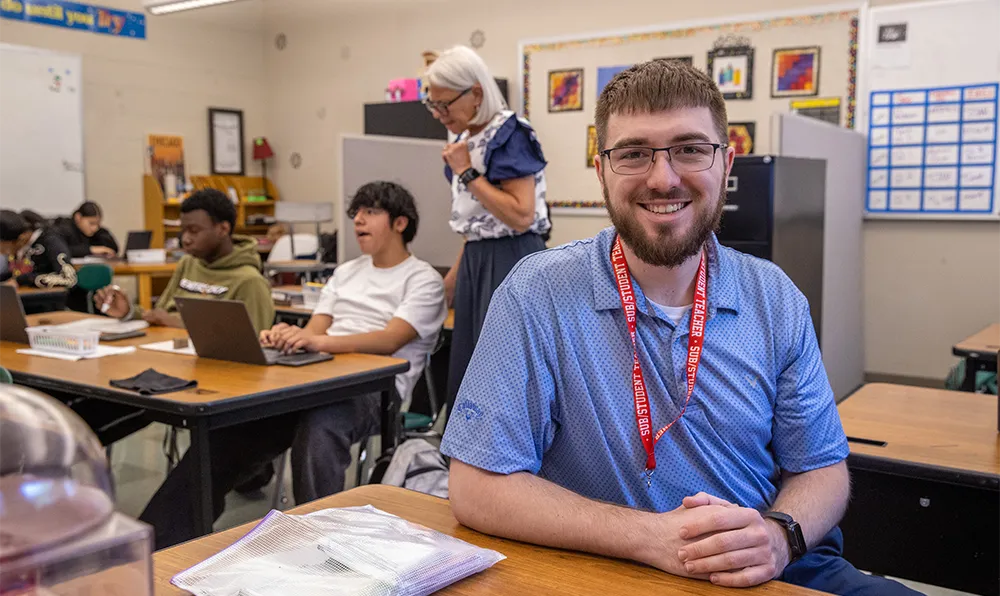 A young man wearing a student teacher lanyard sits at a desk in a high school classroom